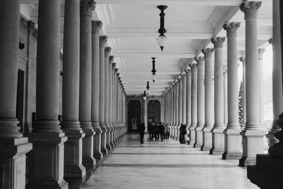 Colunata de um edifício institucional, com as colunas em perspectiva e figuras ao fundo.